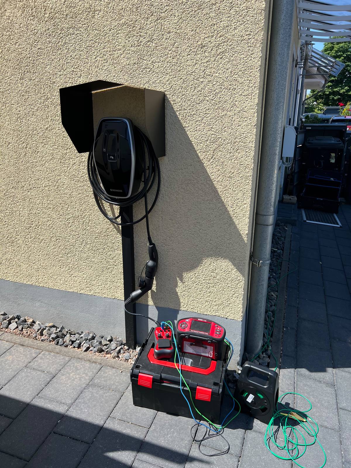 Wallbox mit Wetterschutzdach an einer Hauswand, davor ein Prüfgerät